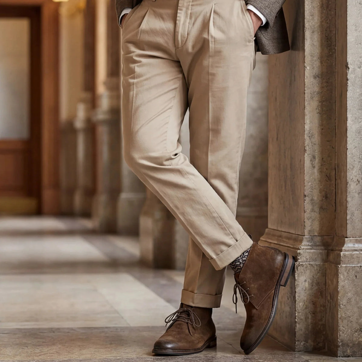 Pampa Suede Chukka Boot with beige pleated trousers, leaning on a column